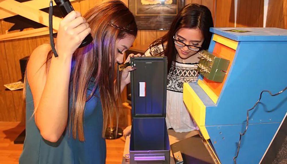 Two individuals are engaged in an interactive science exhibit with one holding an instrument to her head and the other carefully observing a monitor