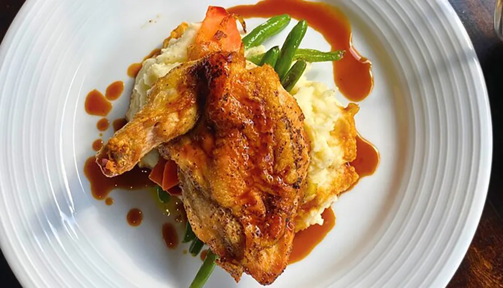A well-presented plate contains roasted chicken on a bed of mashed potatoes accompanied by green beans and carrots drizzled with a brown sauce