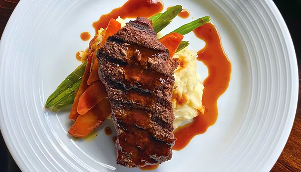 A plate is elegantly presented with a slice of steak a serving of mashed potatoes glazed carrots asparagus and a drizzling of sauce