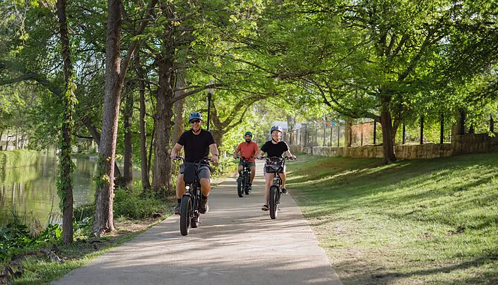 Three individuals are cycling along a scenic paved pathway beside a body of water surrounded by lush green trees