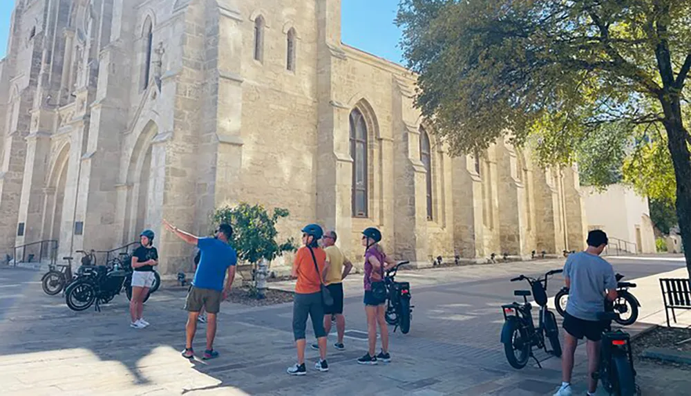 A group of cyclists is gathered near a historical building with one person pointing towards the structure potentially discussing its features