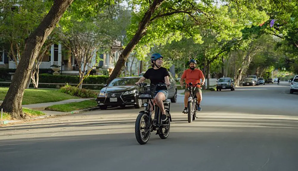 Two people are leisurely riding bicycles on a tree-lined neighborhood street