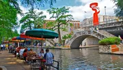 This vibrant image depicts people enjoying outdoor dining by a canal, with colorful umbrellas and a bridge in the background, beneath a large abstract sculpture.