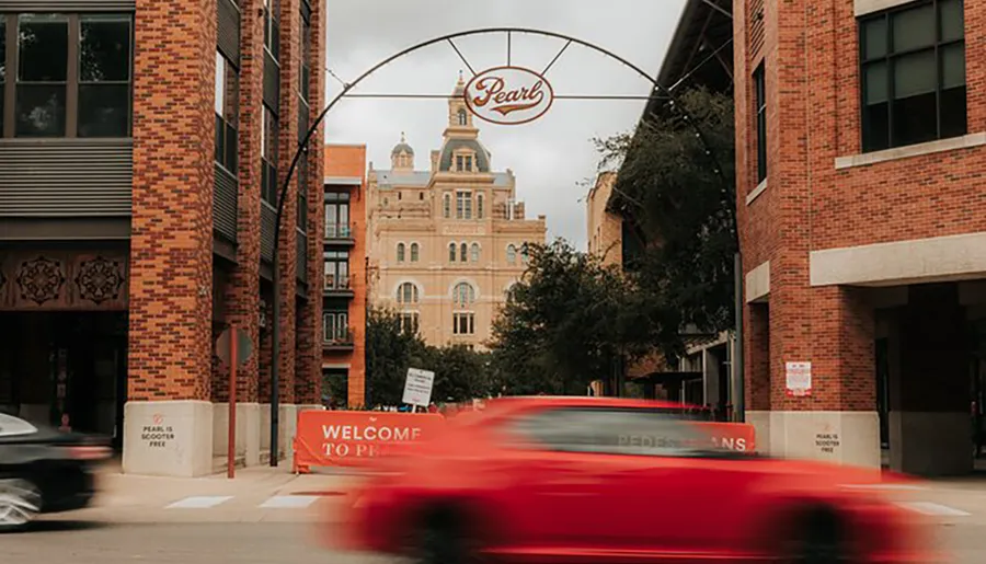 A blurred red car speeds past urban brick buildings under the Pearl archway on a cloudy day.
