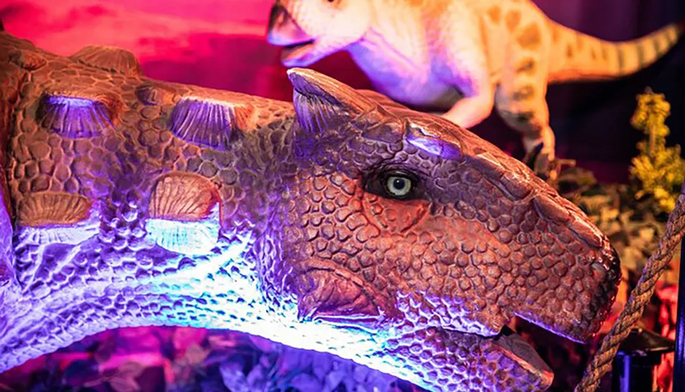 The image shows a close-up of a realistic model of a dinosaur under purple lighting with another dinosaur model visible in the background