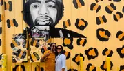 A couple is posing for a photo in front of a vibrant yellow wall featuring an artistic mural with black and orange leopard print and a large portrait.