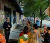 A group of attentive people listen to a speaker during an evening gathering on a city sidewalk lined with trees and storefronts