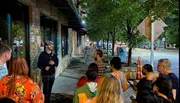 A group of attentive people listen to a speaker during an evening gathering on a city sidewalk lined with trees and storefronts.