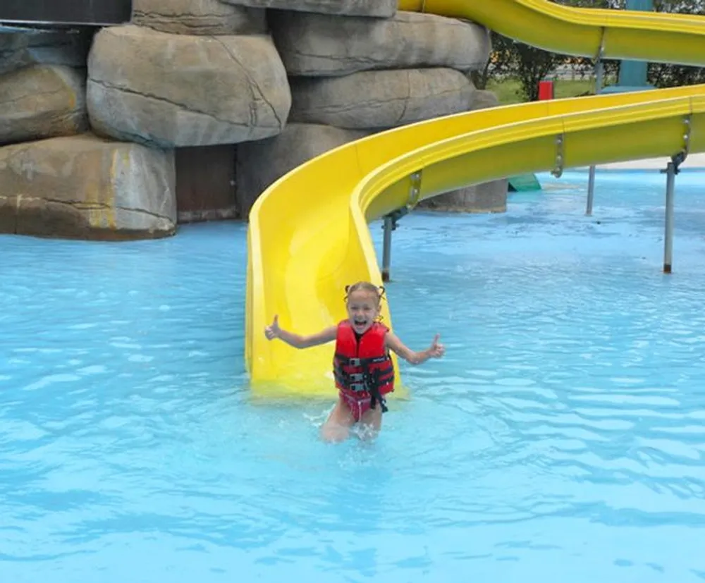 A child with a life vest is gleefully exiting a yellow water slide into a pool