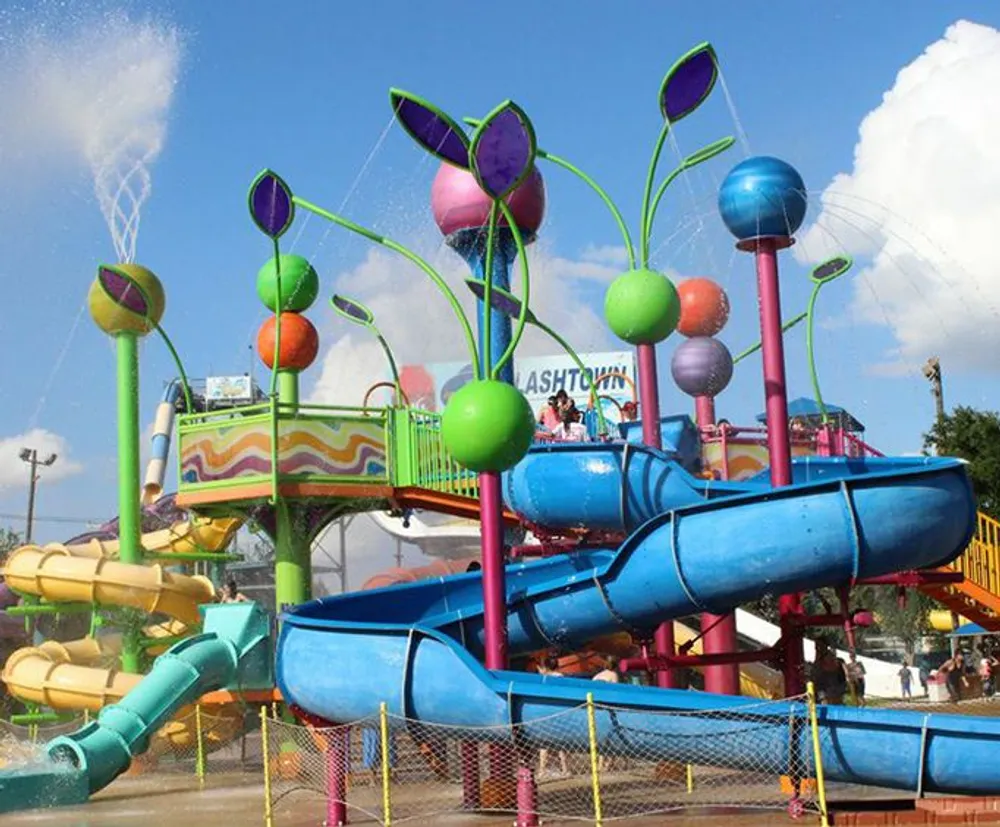 The image shows a colorful water park play structure with several slides and water-spraying features that resemble whimsical flowers