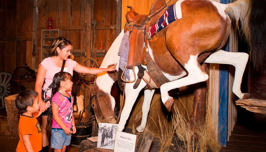 A woman and two children are interacting with a taxidermied horse inside a rustic barn-like setting.