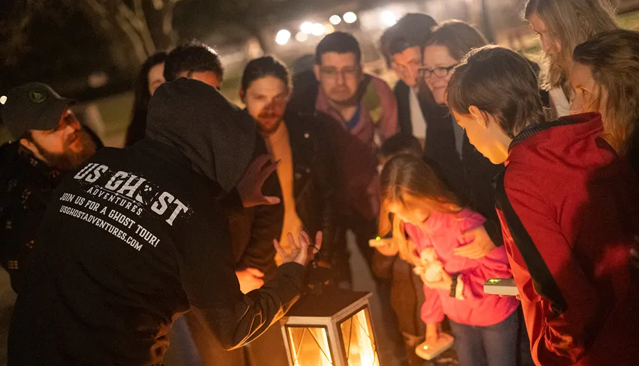A group of people is gathered around a lantern, listening intently to a person where the hoodie indicates they are probably on a ghost tour.