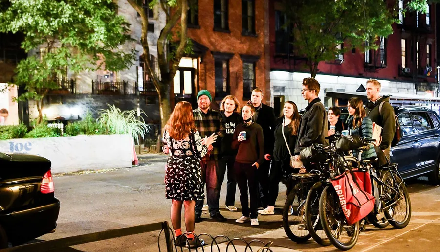 A group of people gather at night on a city street, listening to a woman who seems to be explaining or narrating something to them.