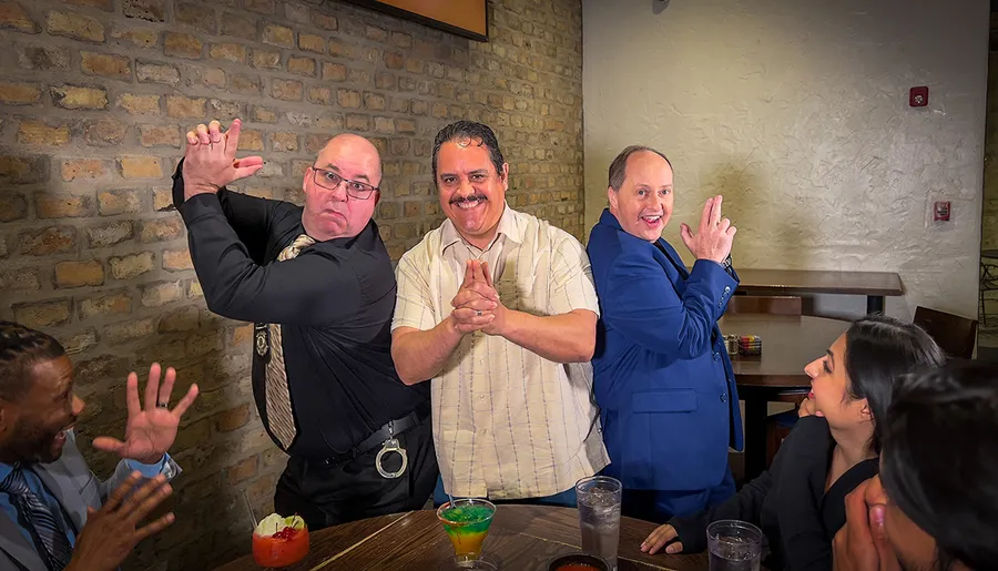 Three men are playfully posing for a photo with exaggerated expressions and hand gestures, while interacting with other people at a casual social gathering.