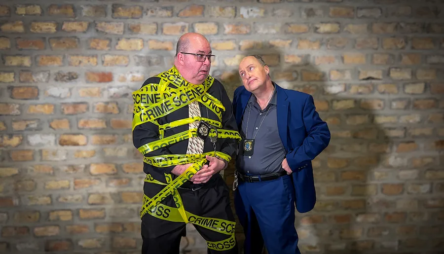 A person wrapped in yellow crime scene tape stands next to another individual posing as a police officer against a brick wall backdrop, creating a humorous scene.