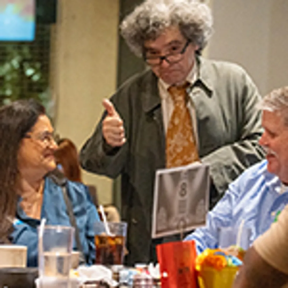 A person with curly hair wearing a coat gives a thumbs-up to two seated individuals at a dining table