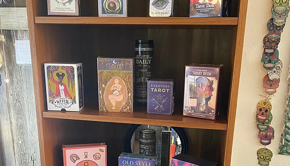 This image features a variety of Tarot card decks displayed on a wooden bookshelf with a string of colorful skull decorations hanging to the right side