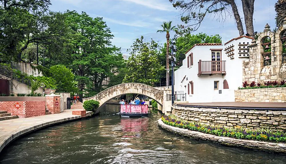 A picturesque scene of a riverwalk with people enjoying a boat tour under an arched bridge with charming white buildings and lush greenery surrounding the waterway