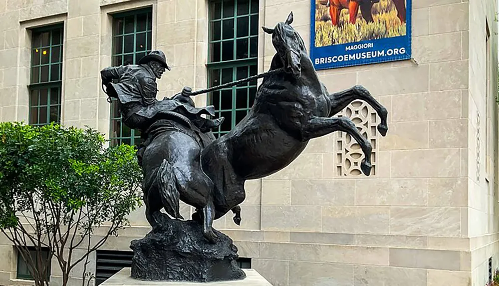 The image shows a dynamic bronze statue of a cowboy on a rearing horse set against the backdrop of a building with stone architecture and greenery