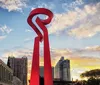 The image shows a large red abstract sculpture under a sky with a colorful sunset with city buildings and a tower in the background