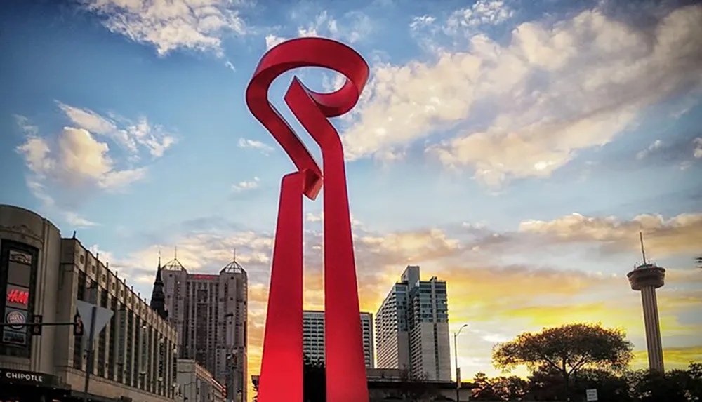 The image shows a large red abstract sculpture under a sky with a colorful sunset with city buildings and a tower in the background