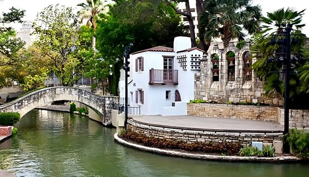 The image shows a picturesque view with a white building lush greenery a footbridge over a canal and architectural elements that suggest a blend of cultures reminiscent of a tranquil urban oasis