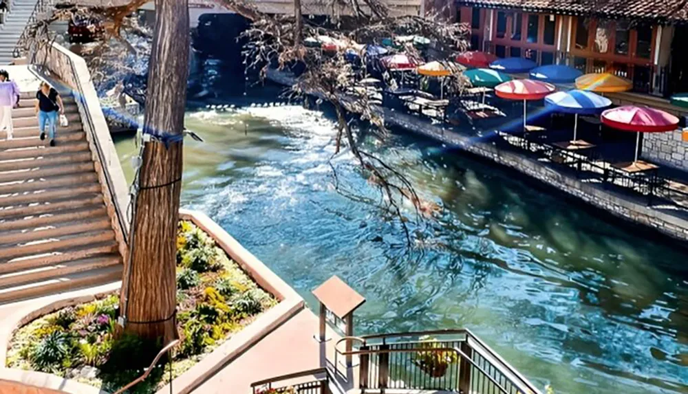 The image shows a sunny outdoor setting with a riverwalk featuring colorful umbrellas at riverside dining areas a staircase leading down to the walkway and people enjoying the ambiance