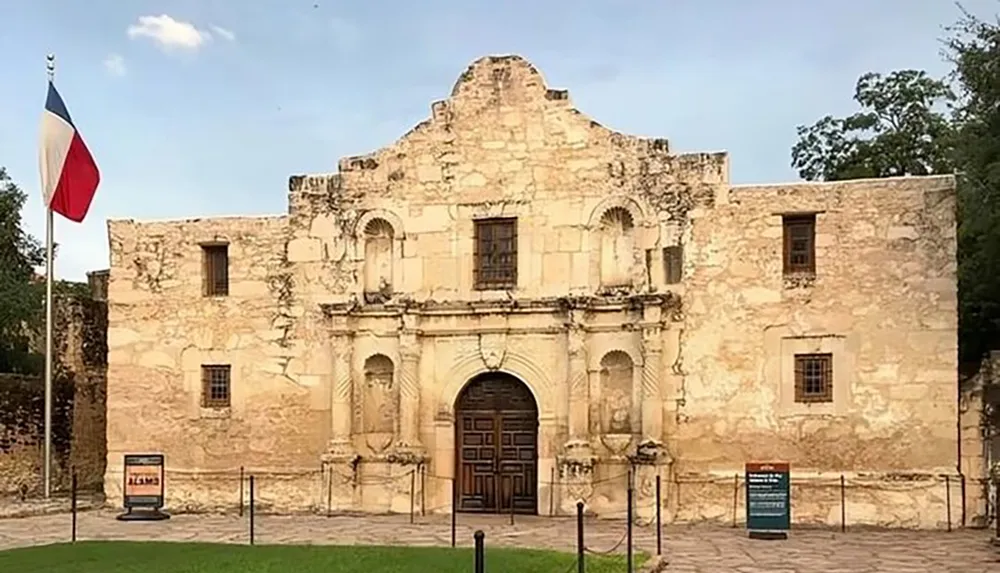 The image shows the historic facade of the Alamo a significant landmark in Texas history with a Texas flag flying on the left