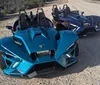 A blue three-wheeled Polaris Slingshot vehicle is parked on the side of a road next to a darker vehicle of the same model