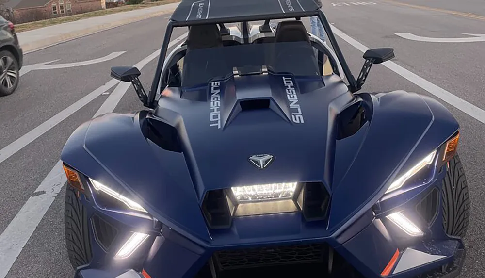 The image shows a blue Polaris Slingshot a three-wheeled vehicle from a frontal perspective on a street during daylight