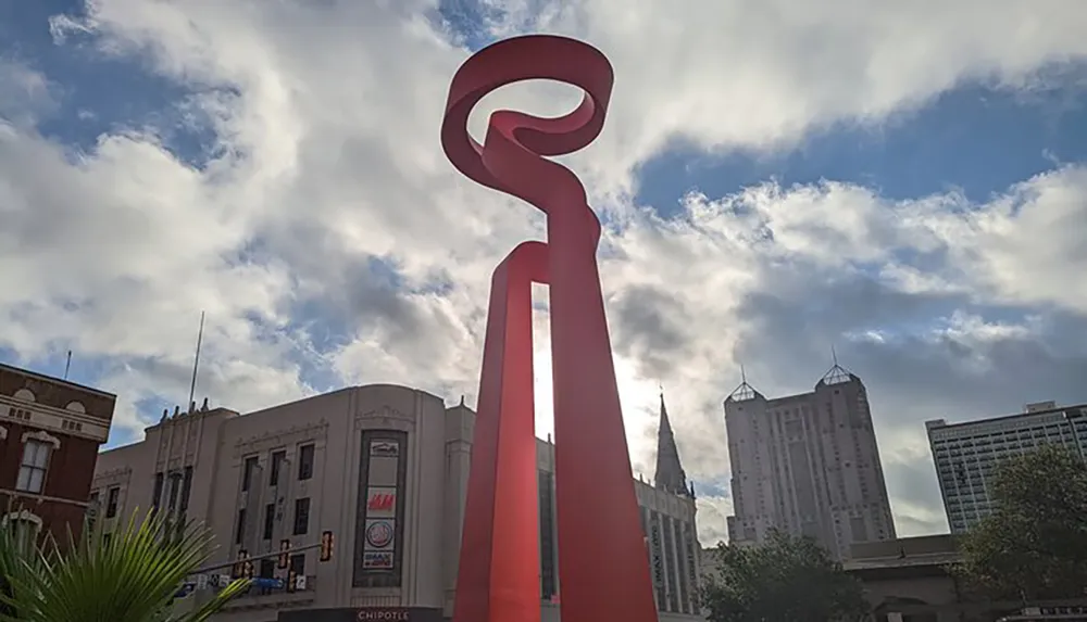 The image shows a large red abstract sculpture set against a blue sky with scattered clouds surrounded by urban buildings