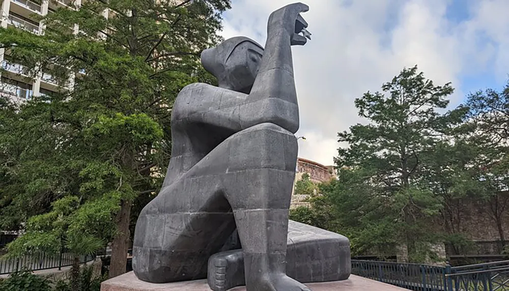 The image shows a large abstract stone sculpture of a seated figure with an outstretched arm against an urban backdrop with trees