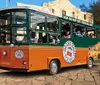 The image shows a colorful sightseeing trolley bus with passengers aboard indicating a hop-on-hop-off tour service under a clear sky with historical architecture in the background