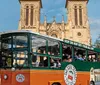 The image shows a colorful sightseeing trolley bus with passengers aboard indicating a hop-on-hop-off tour service under a clear sky with historical architecture in the background