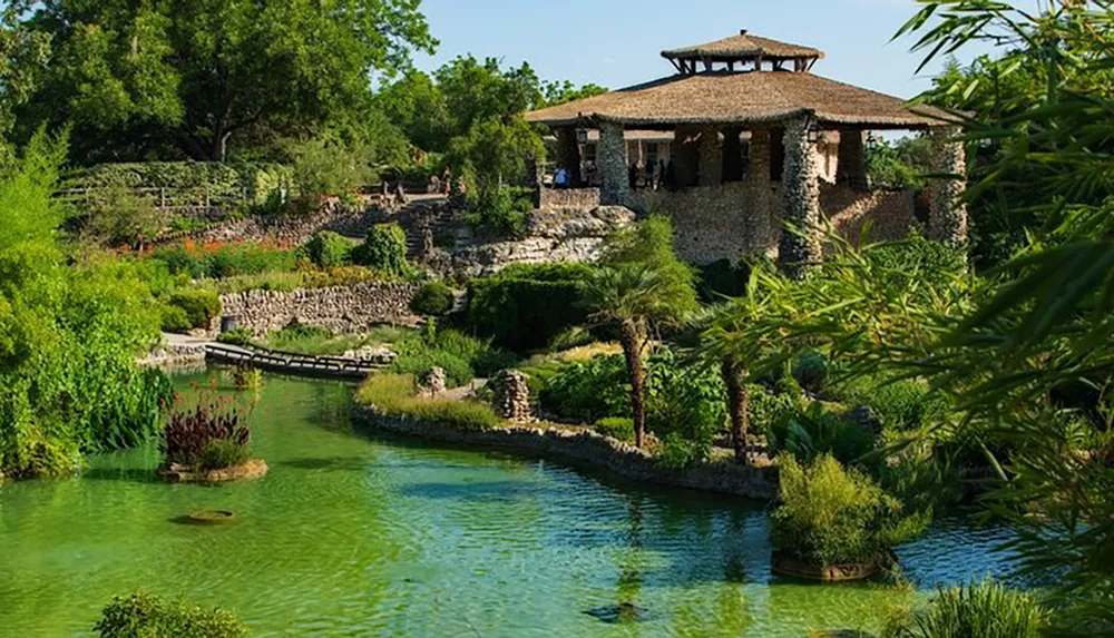 A tranquil scene depicting a lush garden with a clear blue pond a stone bridge and a rustic building surrounded by vibrant greenery under a clear sky