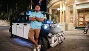 A person is standing confidently with arms crossed next to an electric shuttle vehicle on a city street at night.