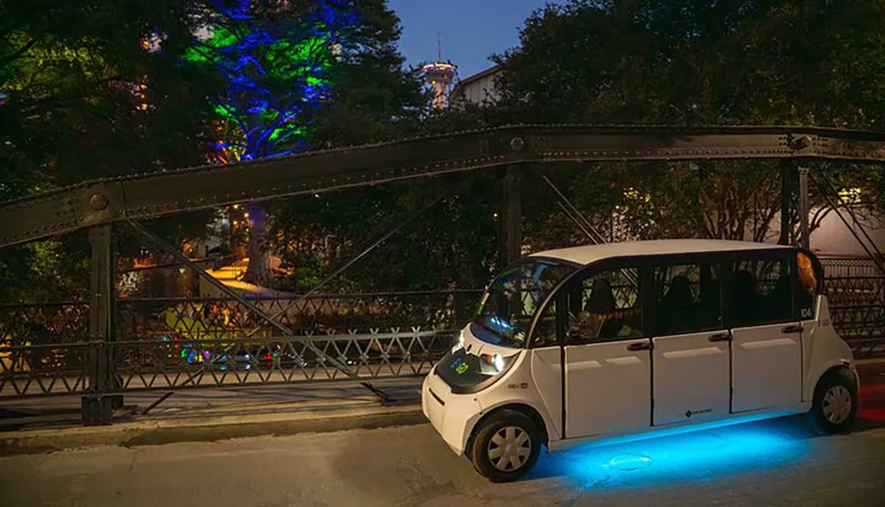 A compact electric vehicle with blue underglow lighting is parked next to an old truss bridge at dusk with trees illuminated by colorful lights in the background