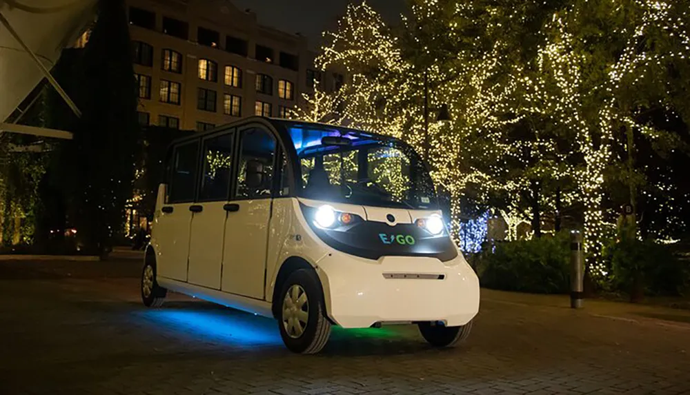 A compact white vehicle adorned with a blue light underglow is parked at night before festively lit trees creating a serene urban scene