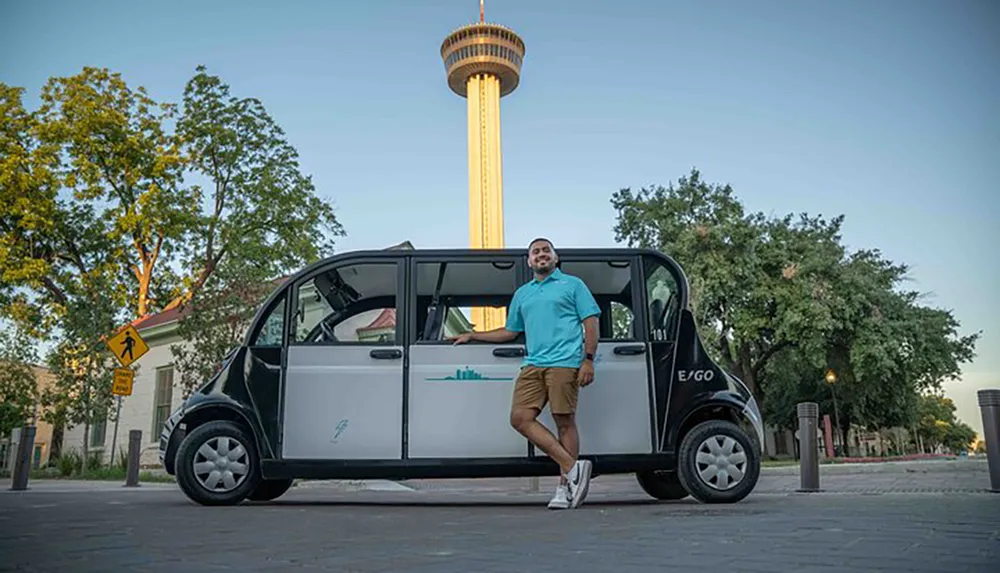 A person is standing with one foot inside a compact electric vehicle smiling towards the camera with a distinctive tower illuminated in the background at dusk