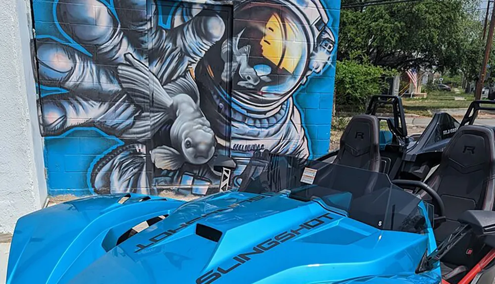 A blue Polaris Slingshot vehicle is parked in front of a vibrant mural depicting an astronaut with a goldfish