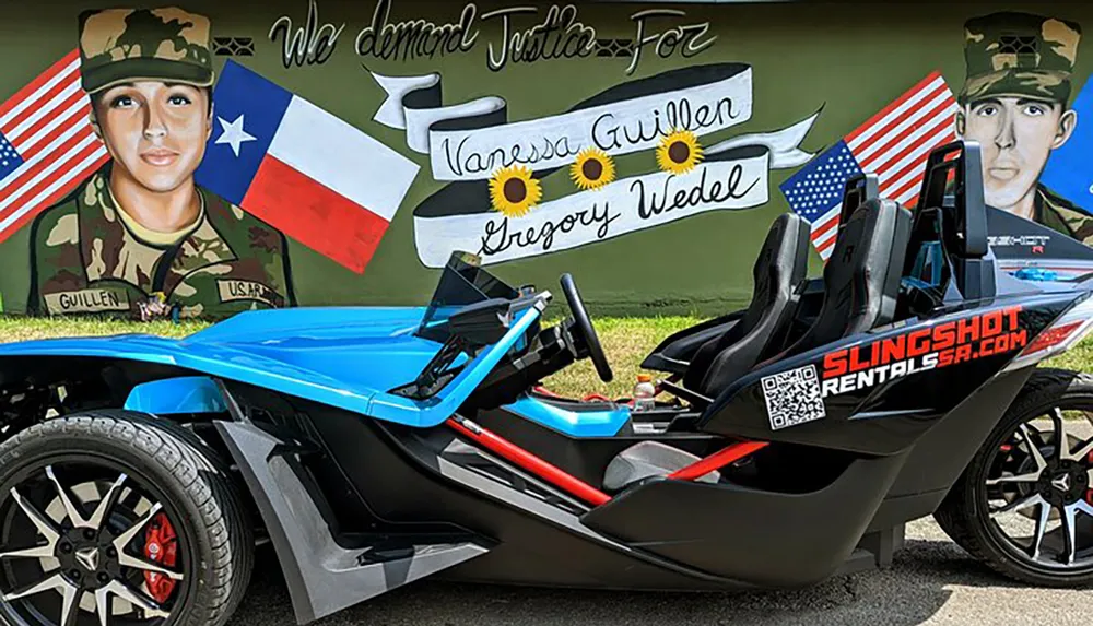 The image depicts a vibrant three-wheeled vehicle a Polaris Slingshot parked in front of a mural paying tribute to two individuals featuring their portraits names and elements of the Texas and United States flags along with sunflowers and a call for justice