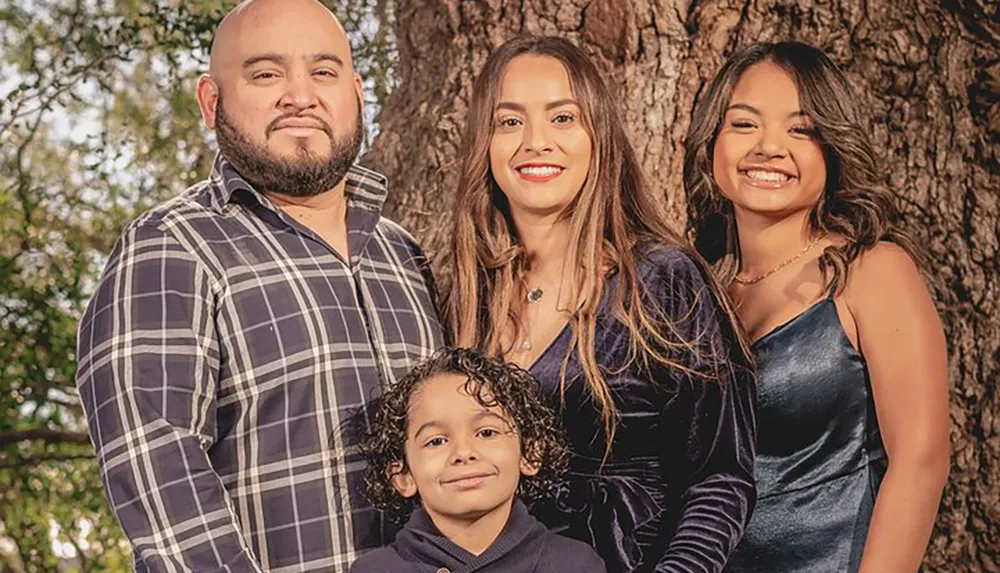 The image shows a family of four smiling for a portrait outdoors with a large tree in the background with two adults flanking a young girl and a small child in front of them