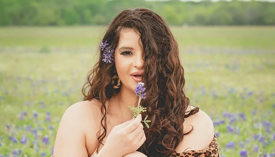 A woman with flowers in her hair is holding a bluebonnet, posing in a field with more bluebonnets in the background.