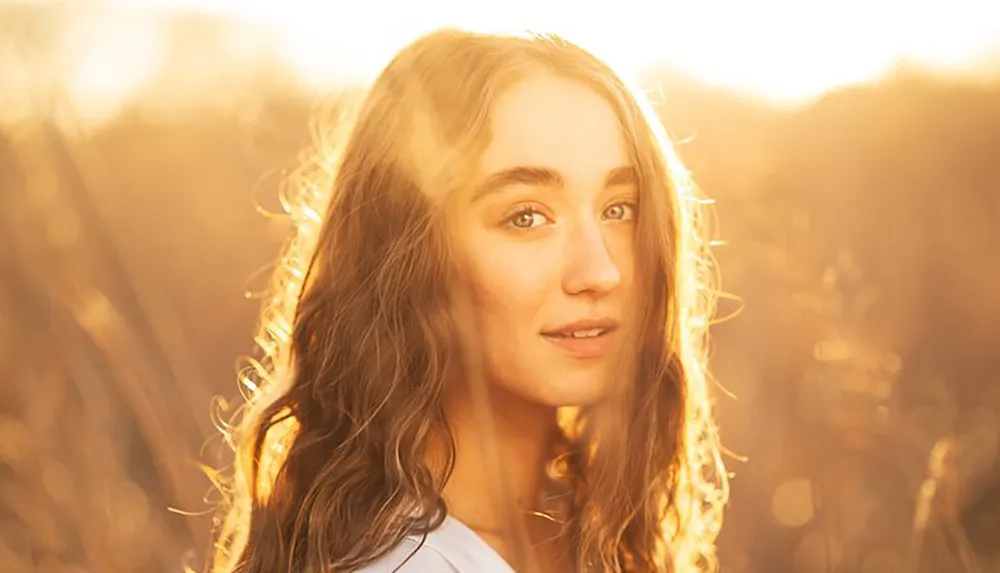 A young woman with wavy hair is backlit by a warm golden sunset creating a soft glowing portrait amidst a blurred natural background