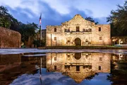 The Alamo Mission in San Antonio, Texas, is illuminated in the early evening and reflected in a smooth water surface in front of it.