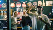 Three friends are enjoying a fun moment in a bar with one of them playfully holding a dart mid-throw, while another holds a cocktail, all surrounded by various bar signage and a person in the background.