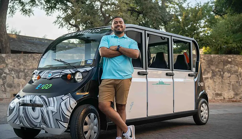 A smiling person with crossed arms is standing proudly next to a compact electric vehicle with sliding doors under a grove of trees