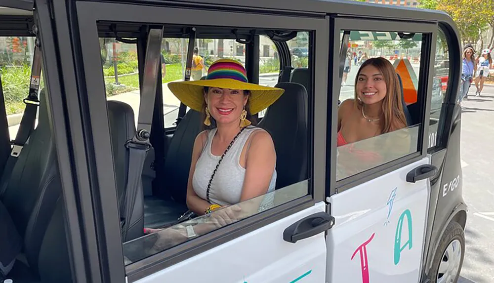 Two smiling individuals are seated inside a small white electric vehicle with an open door the one on the left wearing a colorful wide-brimmed hat