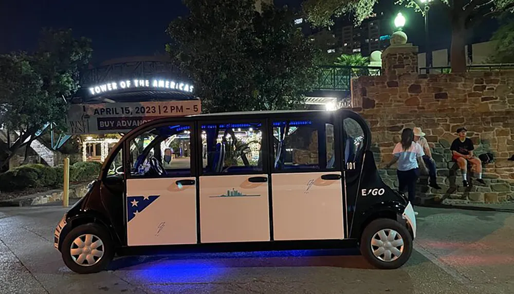 A small electric vehicle adorned with Texas-themed graphics is parked at night with the illuminated Tower of the Americas sign in the background