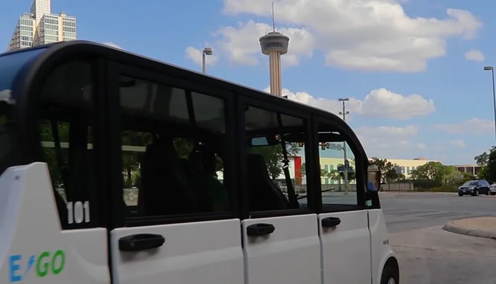 A shuttle van with E GO written on its side is parked with a distinctive tower visible in the background under a blue sky with scattered clouds